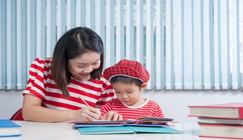 cute-boy-doing-his-school-homework-with-his-mother-home-he-is-writing-book