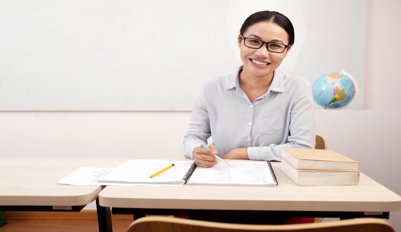 smiling-asian-female-teacher-sitting-desk-classroom-writing-notebook